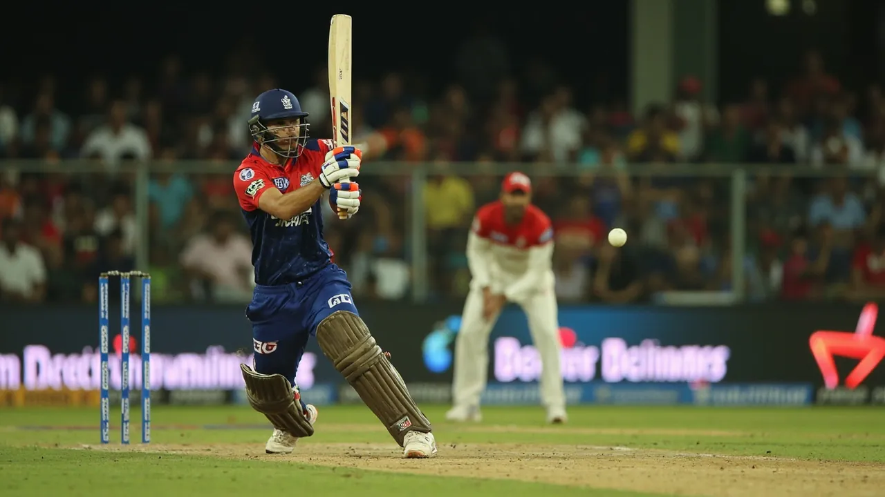 A batsman in mid-stroke during an IPL fixture, follow-through pose with the bat extended as the ball leaves the frame, stands blurred behind